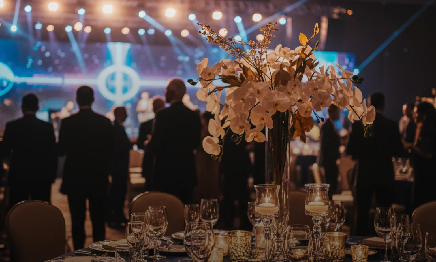 Elegant ballroom event with white floral centerpieces and a blue-lit stage background with guests mingling