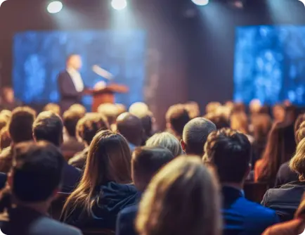 Speaker on stage presenting to a large, attentive conference audience under blue stage lighting